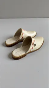 Sandalias cerradas tipo mules de cuero color natural, con detalle de aplique pintado a mano en tonos tierra y herraje metálico triangular en el lateral.