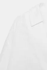 Blusa blanca confeccionada en lino, con cuello polo, escote en V y mangas largas amplias. Presenta un diseño holgado y bajo asimétrico.