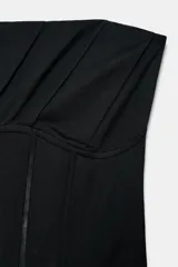 Corsé de escote strapless con detalle de drapeado en el pecho y varillas frontales visibles. Presenta un bajo asimétrico y cierre en la espalda.