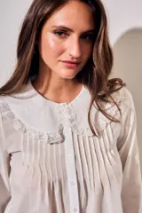 Blusa blanca de algodón peinado con mangas largas, cuello babero bordado y pliegues frontales.