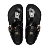 Sandalia tipo ojota Birkenstock modelo Gizeh con diseño de tira de dedo, confeccionada en cuero negro con acabado brillante. Presenta una hebilla grande de metal dorado y plantilla anatómica de corcho y látex con suela de goma.