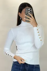 Polera de hilo blanca de cuello alto y manga larga, con detalle de botones dorados en los puños. Tejido de punto acanalado ajustado al cuerpo.