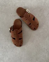 Zueco tipo sandalia de estilo jaula, confeccionado en gamuza negra con tiras entrelazadas y hebilla metálica dorada lateral. Cuenta con plantilla acolchada con logo de la marca.