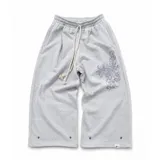 Pantalón de jogging gris de corte baggy, confeccionado en algodón. Presenta cintura elástica con cordón largo ajustable, botones en la parte inferior para ajustar la botamanga y un diseño artístico que combina estampa en plastisol con bordado en relieve sobre el muslo izquierdo.
