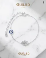 Pulsera de plata con cadena eslabonada, dije central en forma de mano de Fátima (Hamsa) recubierto de circonias, y un dije colgante circular con diseño de ojo turco con circonias azules y blancas. Cuenta con cadena de alargue y cierre de resorte.