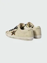 Championes Golden Goose modelo Super Star, con diseño de efecto desgastado. Presentan cuerpo en cuero color crema, puntera y lengüeta con acabado de glitter dorado, estrella lateral con estampado animal print y cordones negros.