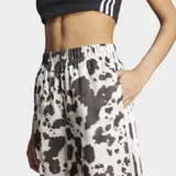 Pantalón Adidas de satén con estampado animal print tipo vaca en tonos blanco y negro, de corte holgado, perneras anchas y fluidas, cintura elástica y las icónicas 3 tiras de la marca en los laterales.
