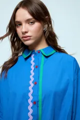 Camisa azul de corte oversize, con cierre frontal de botones y detalles de cintas onduladas en colores contrastantes a lo largo de la botonadura y en los puños. Presenta cuello clásico y mangas largas.