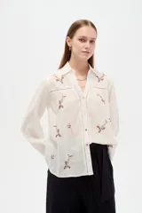 Camisa de algodón blanca de corte holgado, con cuello camisero, mangas largas y bordados florales multicolores. Presenta detalles de calados en los hombros y cierre frontal con botones.