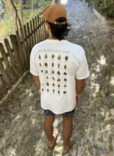 Remera blanca de algodón con cuello redondo y estampado en la espalda con imágenes de cogollos de marihuana y texto "BROKERS HIGH QUALITY GOODS".