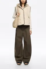 Chaleco acolchado color crema con capucha integrada, cierre frontal de botones a presión y vivos en contraste color negro. Presenta diseño de costuras geométricas y bolsillos laterales.