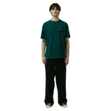 Remera de manga corta color verde oscuro, con cuello redondo y un pequeño parche cuadrado negro con texto morado en el pecho izquierdo.