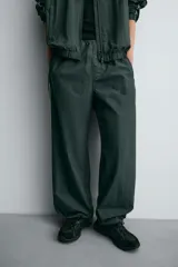 Pantalón jogger de corte relajado confeccionado en tejido técnico de color verde oscuro. Presenta cintura elástica con cordón interior, bolsillos laterales con cierre y bajo ajustable con elástico. Incluye detalles de líneas en contraste a lo largo de las piernas.