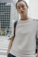 Top de cuello redondo y manga corta con diseño de capas que combina una remera de algodón blanca con un chaleco de punto gris superpuesto. Presenta detalles de botones joya en el hombro.