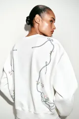 Sudadera de algodón con cuello redondo y manga larga. Presenta un diseño minimalista en color blanco con un delicado bordado de un lazo con flores en el pecho.