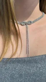 Gargantilla tipo choker de metal plateado con diseño de lazo central decorado con cristales brillantes y dos tiras colgantes finas también cubiertas de cristales.