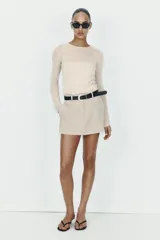 Falda pantalón corta de tiro medio en color beige, con bolsillos delanteros y cierre frontal de cremallera. Incluye un cinturón negro integrado con hebilla metálica redonda.