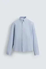 Camisa de algodón de corte relajado, con cuello solapa abotonado (button-down) y manga larga terminada en puño con botón. Presenta un patrón de rayas verticales finas en tonos azul claro y blanco.