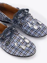 Mocasín de tela con estampado de cuadros en tonos azules y marrones, con detalles de cuero liso, cordones decorativos y apliques metálicos en la parte frontal.