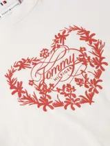 Remera de algodón de corte regular, color blanco, con cuello redondo y mangas cortas. Presenta un estampado frontal en color rojo que combina un diseño floral en forma de corazón con el logo de la marca en el centro. Incluye el logo de la marca bordado en la manga.