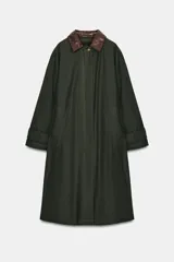 Trench oversize de color verde oscuro, con cuello de piel en tono marrón a contraste. Presenta manga larga con trabillas en los puños, bolsillos delanteros, cierre frontal con cremallera y botones ocultos bajo solapa, y abertura en la espalda.