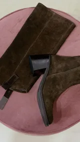 Bota de caña alta de gamuza en color marrón chocolate, con taco cuadrado y puntera cuadrada. Cuenta con un diseño holgado en la caña y detalle de trabilla decorativa en el tobillo.