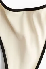 Bombacha de bikini vedetina de tiro bajo color marfil, con bordes ribeteados en negro y cobertura media en la parte trasera.