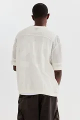 Remera de malla color blanco hueso, de manga corta, con cuello redondo y hombros caídos. Presenta un diseño de tejido perforado con un número grande grabado en el frente.