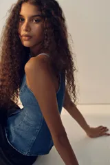 Chaleco sastrero en denim de algodón rígido, con escote recto, breteles finos y cierre frontal de botones metálicos. Presenta costuras princesa al frente y una abertura pronunciada en el dobladillo inferior.