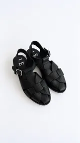 Sandalias de cuero negro con diseño de tiras trenzadas artesanales en el empeine, tira ajustable al tobillo con hebilla metálica y suela plana.