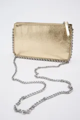 Cartera pequeña color dorado metálico con cierre superior. Presenta tachas plateadas decorativas a lo largo de los bordes superior e inferior.