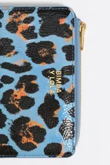 Billetera mediana con exterior rígido y textura brillante, con estampado de leopardo en tonos azul celeste, negro y naranja. Presenta cierre de cremallera y logo metálico de Bimba y Lola en el frente.