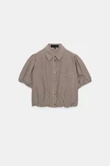 Camisa corta de cuello solapa y manga corta abullonada acabada en elástico, color verde con estampado de cuadros vichy. Cuenta con un bolsillo de plastrón en el delantero, bajo con elástico y cierre frontal con botones.