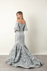 Vestido largo de fiesta color gris perla, corte sirena, con escote off-shoulder y mangas cortas con volados. Confeccionado en tul bordado con hilo al tono y leve brillo.