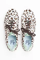 Zapatillas de estilo running con estampado animal print de leopardo sobre fondo blanco. Presentan detalles en piel verde tornasolada en la parte trasera y lengüeta, cordones oscuros y suela de goma dentada con cuña interna.