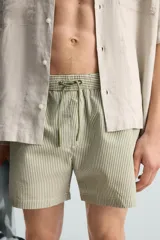 Bañador de hombre color verde claro con finas rayas blancas verticales (efecto seersucker). Tiene cintura elástica ajustable con cordón verde, bolsillos laterales y un bolsillo trasero de vivo.