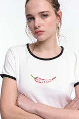 Remera baby blanca de algodón, de corte cropped al cuerpo y mangas cortas. Presenta ribetes negros en contraste en el cuello redondo y los puños, con un estampado localizado de un ají rojo con la inscripción 'Pimentón' en el centro del pecho.