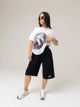 Remera blanca de manga corta de la colección Tini x Under Armour, con cuello redondo y diseño holgado. Cuenta con un gran estampado fotográfico en el centro y una etiqueta negra con logotipos en el borde inferior.