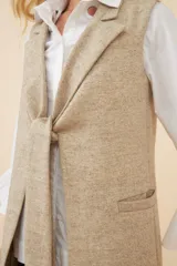 Chaleco largo color marrón claro, abierto, con lazo en la delantera y bolsillos ojal. Confeccionado en lana fina sin teñir.