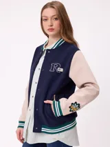 Campera estilo varsity con cuerpo en color marrón y mangas en tono beige. Presenta cierre frontal con botones a presión, cuello, puños y ruedo con franjas tejidas en verde y blanco. Incluye un parche con la letra R en el pecho y detalles bordados en las mangas.