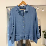 Camisa azul jean de manga larga y corte oversize, con cierre frontal de botones y detalles de volados o pliegues superpuestos en el ruedo y en los puños.