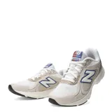 Championes urbanos New Balance modelo Amaste, con diseño en tonos beige y blanco, detalles en azul y rojo en el logo lateral y la lengüeta, confeccionados en malla y materiales sintéticos con suela de goma.