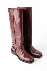 Botas de cuero croco color rojo oscuro, de caña alta (38 cm), con taco bajo de 3 cm y punta cuadrada. Presentan un arnés decorativo con hebilla metálica alrededor del tobillo.