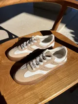 Zapatillas urbanas de cuero color beige y blanco, con tachas plateadas en los laterales y suela de goma color caramelo. Diseñadas en Uruguay y producidas en Brasil.