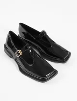 Mocasín estilo T-bar color negro en charol, con punta cuadrada, tira en el empeine con hebilla dorada y suela plana.