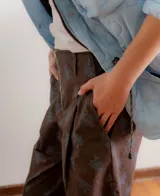 Pantalón sastrero de tiro medio con cierre frontal visto y bolsillos laterales. Presenta un diseño de corte recto y amplio con estampado floral sutil sobre fondo marrón.