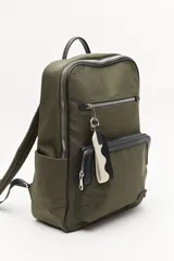 Mochila urbana de nylon en color verde militar, con detalles en cuero sintético negro. Cuenta con compartimento principal con cierre, bolsillo frontal con cierre, bolsillos laterales abiertos y asa superior. Incluye un colgante decorativo de tela y correas ajustables acolchadas.