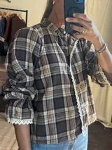 Camisa de franela a cuadros en tonos marrones y beige, con cuello mao, cierre frontal de botones y detalles de puntilla blanca en la tapeta y los puños. Presenta mangas con volumen y puños elásticos.