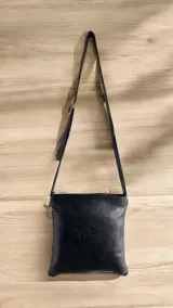 Bandolera negra de cuero con solapa frontal decorada con una intervención artística pintada a mano y correa larga ajustable para llevar al hombro o cruzada.