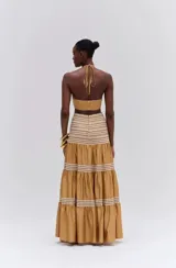 Vestido largo color mostaza, con diseño cut-out en la cintura, cuello halter con tiras para atar, y detalles bordados en el escote. La falda larga presenta paneles fruncidos con detalles tejidos en hilo crudo.